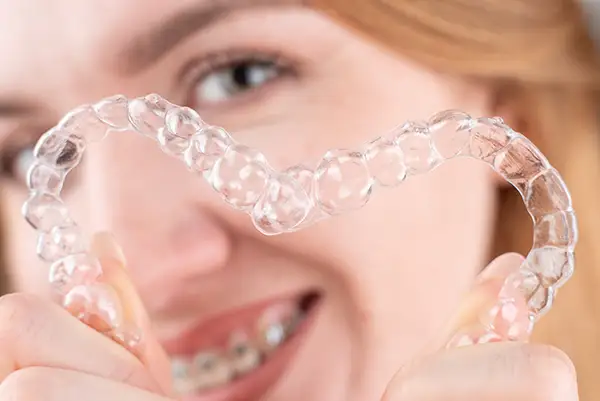 Smiling woman with braces holding clear aligners in a heart shape, showcasing orthodontic solutions. Smiling woman with braces holding clear aligners in a heart shape, showcasing orthodontic solutions.