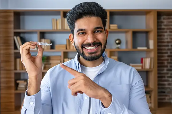 Smiling man holding and pointing at an Invisalign clear aligner in a home setting, highlighting its use for adult teeth alignment. Smiling man holding and pointing at an Invisalign clear aligner in a home setting, highlighting its use for adult teeth alignment.