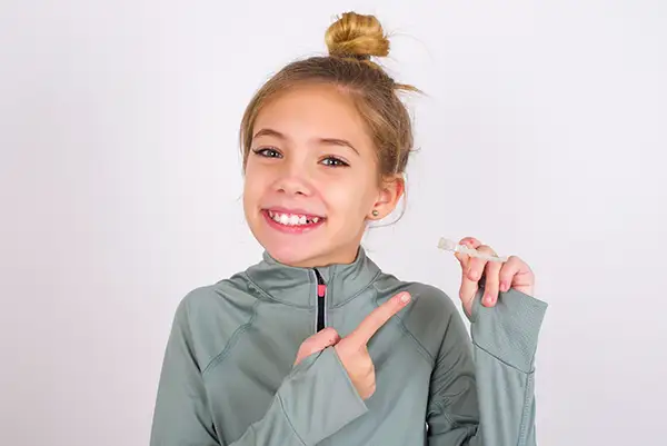 Young girl in a gray sweatshirt smiling and pointing to an Invisalign clear aligner she is holding, promoting orthodontic care for teens. Young girl in a gray sweatshirt smiling and pointing to an Invisalign clear aligner she is holding, promoting orthodontic care for teens.