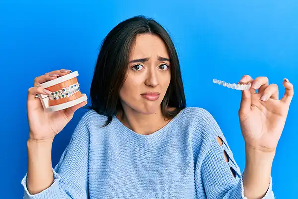 A confused young woman, uncertain about which orthodontic treatment is right for her, holds up clear aligners in one hand and a dental model will traditional braces in the other hand. A confused young woman, uncertain about which orthodontic treatment is right for her, holds up clear aligners in one hand and a dental model will traditional braces in the other hand.