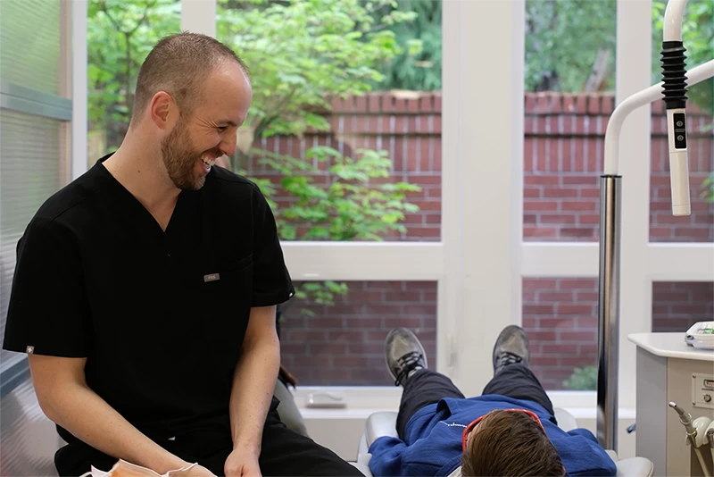 Dr. Christensen smiling and talking to a patient who is lying on a dental chair for his dental exam. Dr. Christensen smiling and talking to a patient who is lying on a dental chair for his dental exam.